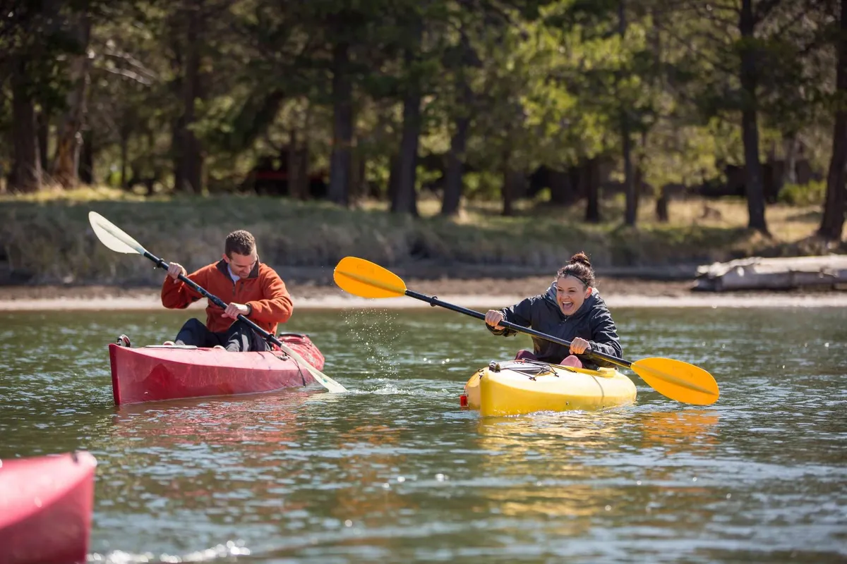 Participants paddle red and yellow kayaks on a calm lake, the water reflecting the vibrant colors and surrounding nature. The scene is active, joyful, and outdoors-oriented, highlighting another type of gear (paddleboards/kayaks) mentioned in the section.