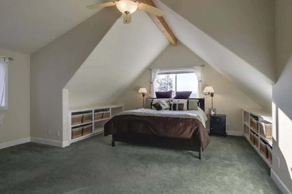 A warmly lit bedroom featuring a comfortable, made-up bed beside a window with bookshelves. Soft natural light filters in, highlighting the room’s cozy and inviting atmosphere—perfect for resting, reading, or starting the day peacefully.