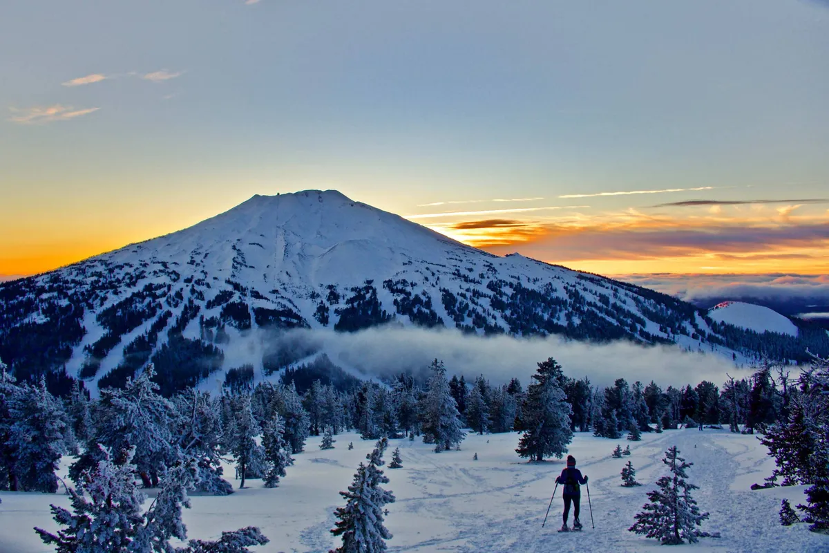 A breathtaking mountain view with a snow-covered peak rising above a dense tree line, a lone hiker, and a sunset sky. The scene captures a sense of adventure and tranquility, evoking opportunities for winter sports like skiing and snowshoeing as well as serene moments in nature.
