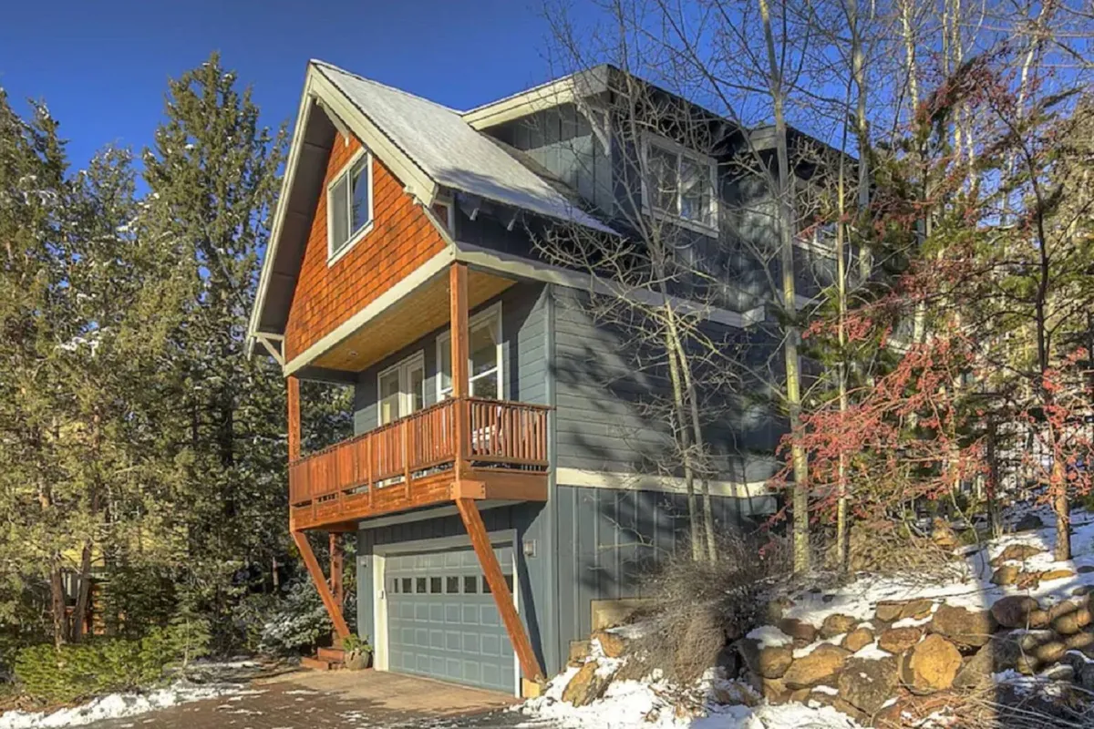 The image showcases the inviting exterior of Quincy House, a single-family home in Bend, Oregon, featuring a charming wooden balcony overlooking a professionally landscaped yard with stonework, framed by graceful aspen trees and a picturesque blanket of snow. The setting evokes a tranquil, cozy atmosphere ideal for relaxing or entertaining outdoors year-round.