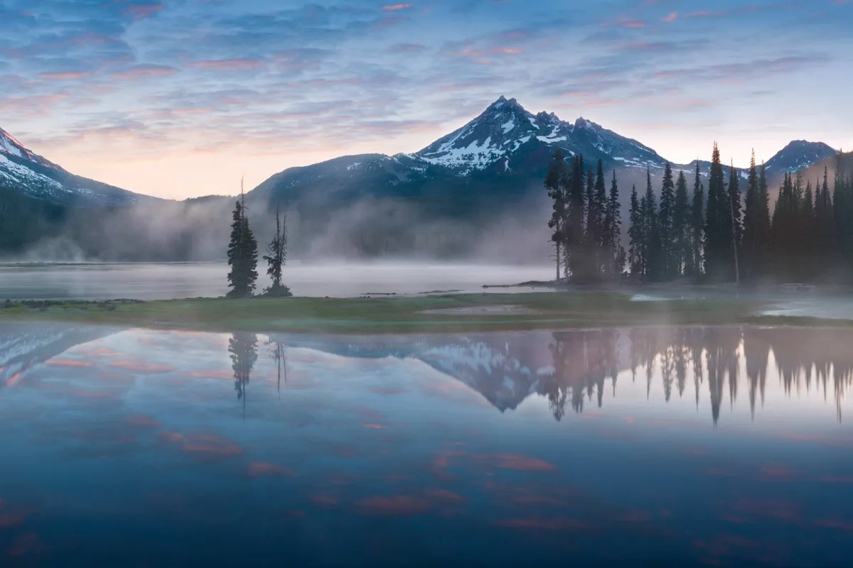 A sweeping view of Paulina Mountain rising over a tranquil lake, framed by lush trees and a soft morning fog. The broad sky overhead casts a gentle light on the landscape, inviting a sense of serenity and escape.
