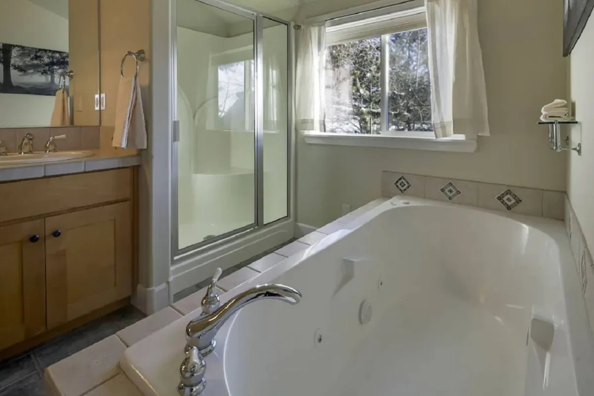 A serene bathroom setting featuring a soaking bathtub positioned near a window with a view of nature. Soft, natural light fills the space, and the shower area is visible, reinforcing an atmosphere of calm and relaxation.