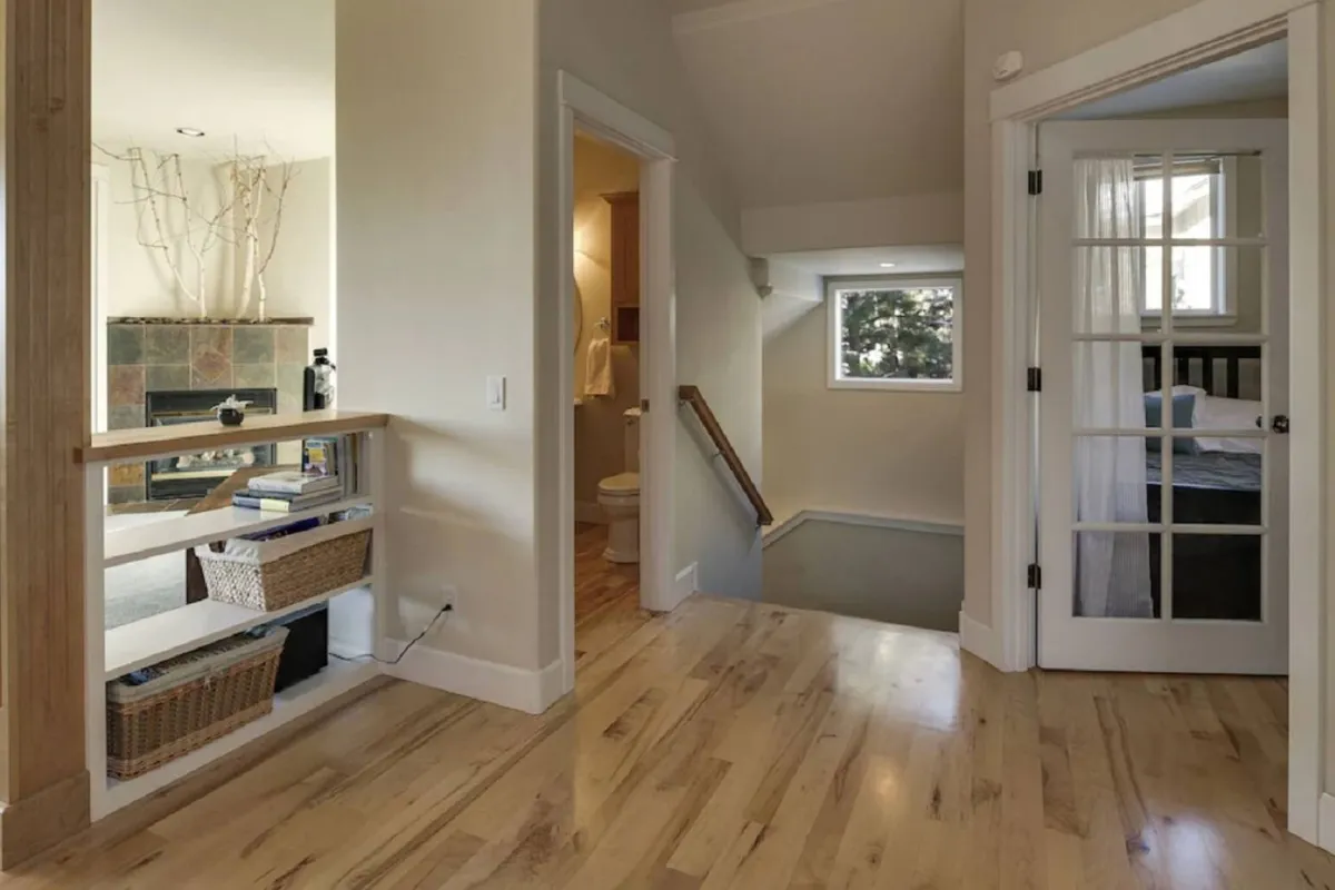 A welcoming great room featuring a cozy fireplace, wooden staircase, and soft sunlight pouring in. The layout suggests an open, airy space leading to both relaxation and intimate gatherings, echoing the comforts of a private retreat.