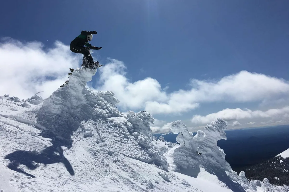 A dynamic action shot of a snowboarder mid-air against a backdrop of a snow-covered mountain peak and cloudy sky. The image radiates energy, excitement, and the thrill of winter sports.