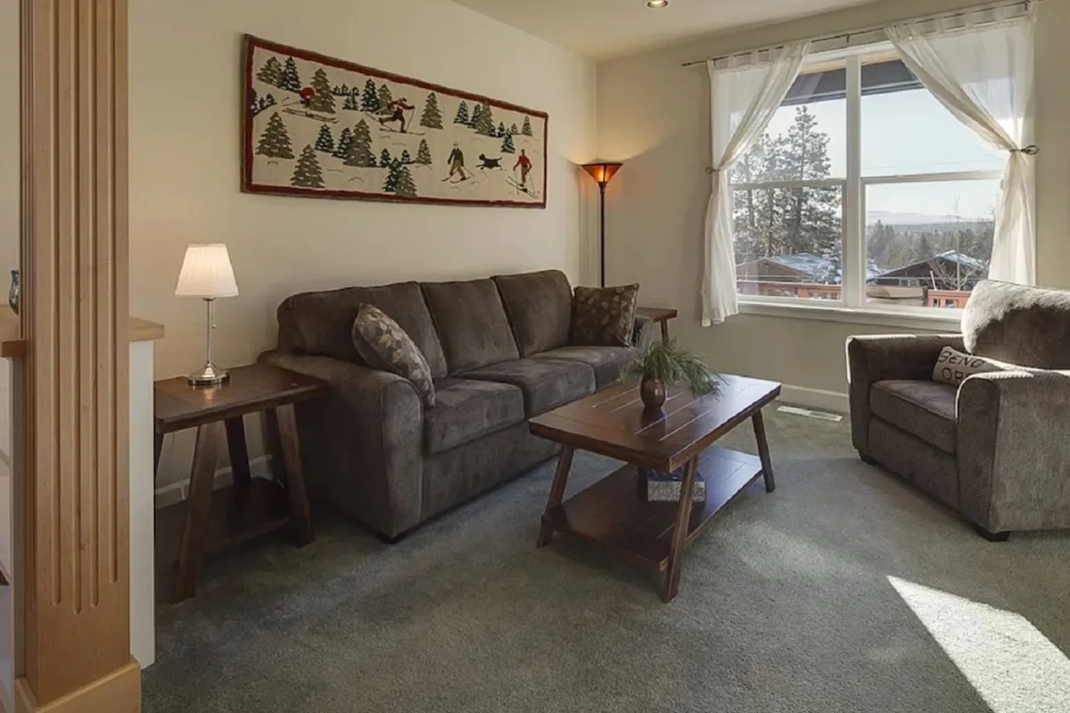 A sunlit great room centered around a plush sofa and coffee table, with large picture windows framing views of the outdoors. The space features warm wall art and is styled for both relaxation and socializing, with ample seating and natural light pouring in.