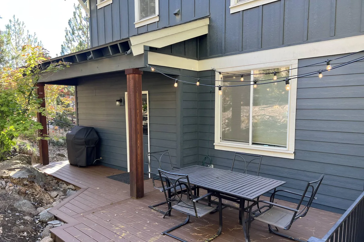 A casual and inviting back patio scene featuring an outdoor dining table beneath string lights, with a new grill (Weber BBQ) positioned nearby. The setup hints at lively barbecues and relaxed al fresco meals, while the fenced backdrop ensures privacy and safety for families.