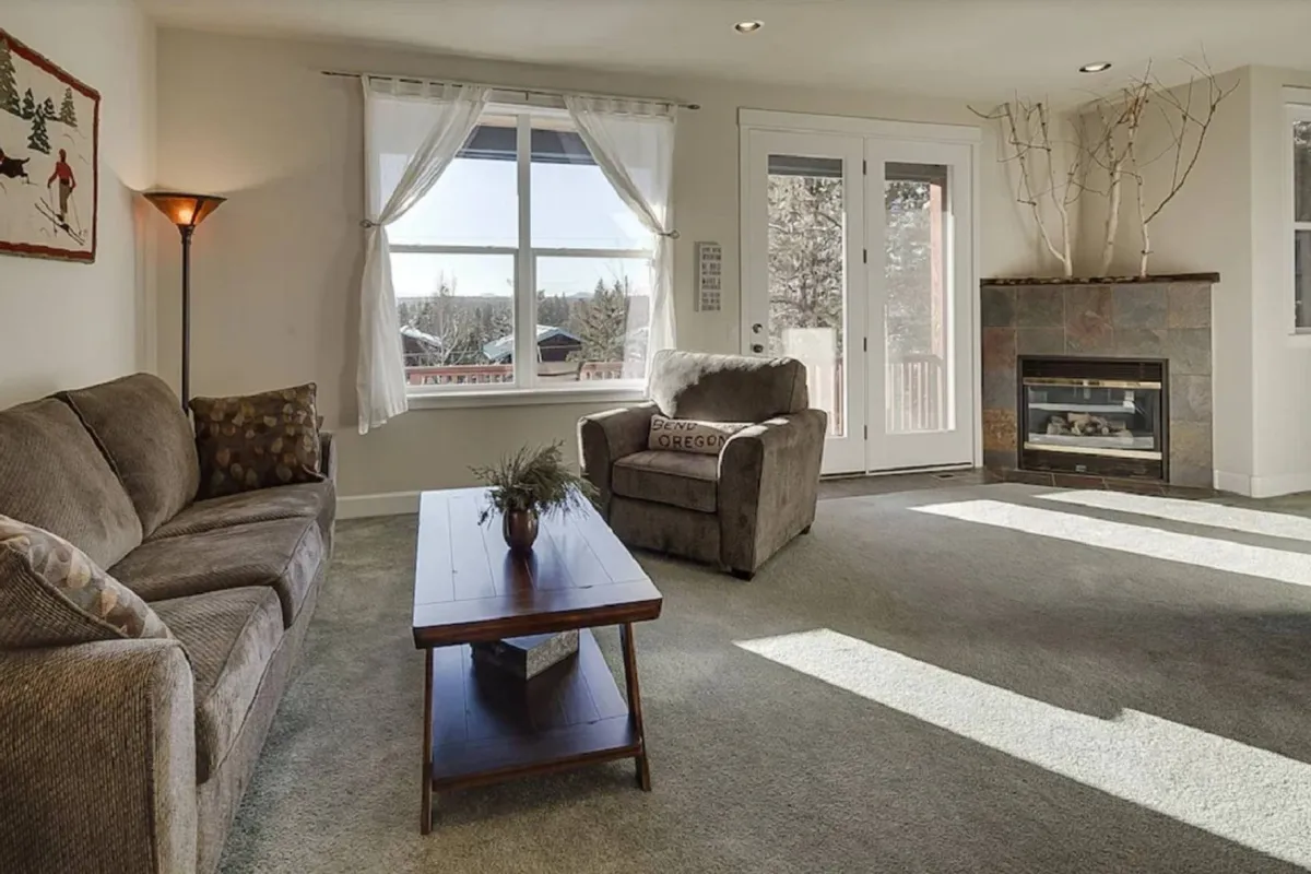 A bright living room featuring a comfortable sofa, a central coffee table, a warm fireplace, and a large window that brings in plenty of natural light. The room feels open and welcoming, suggesting a space ideal for socializing or relaxing after a day of exploring the neighborhood.