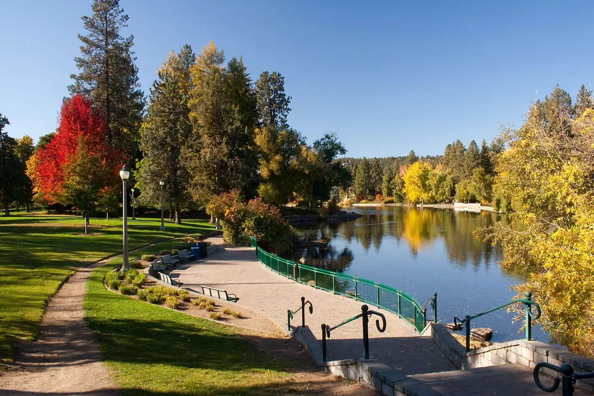 A scenic riverside view showing a calm water surface bordered by colorful trees, a walking path with green railings, and park benches. The area exudes tranquility and natural beauty, perfect for outdoor activities like strolling, picnicking, fishing, or bird watching.