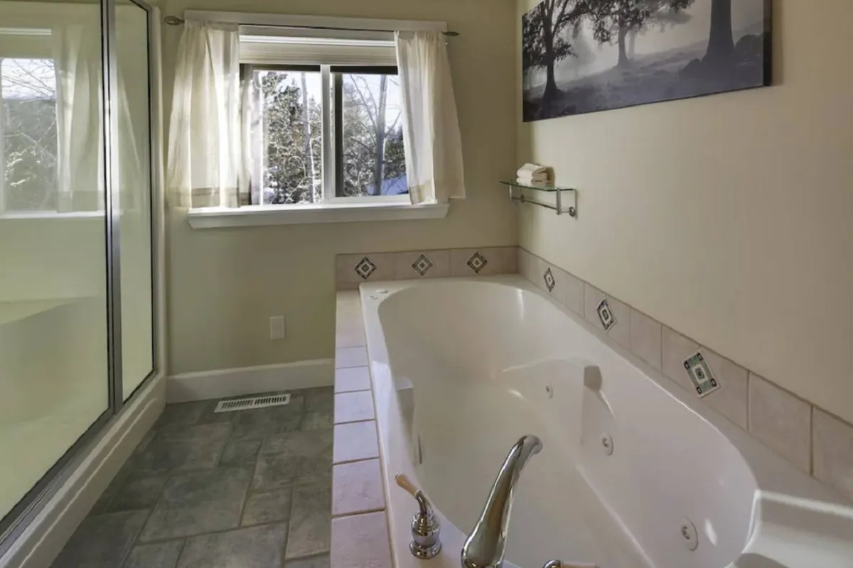 A spa-inspired bathroom featuring a jetted bathtub set beneath a window, a glass-enclosed rainshower, and tasteful decorative art. The scene is bathed in peaceful, natural light, emphasizing relaxation and luxury.