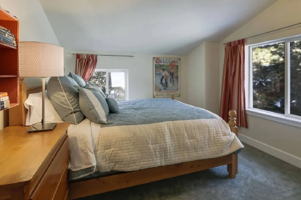 A serene bedroom with a neatly arranged bed with blue bedding, a spacious window framed by curtains, a wooden dresser, and a soft-glowing table lamp. The scene conveys a peaceful, restful retreat enhanced by views and ample daylight.