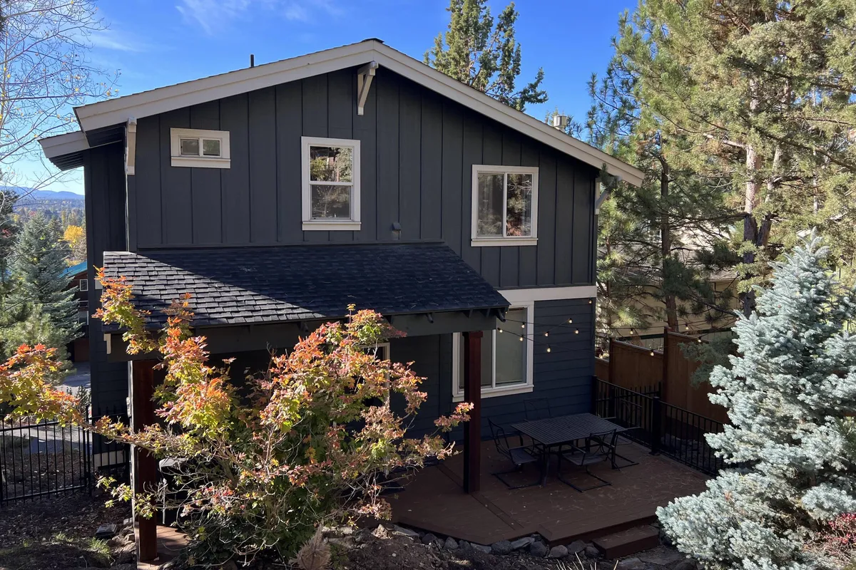 A broad exterior view showing the house’s inviting outdoor seating area surrounded by mature trees, with comfortable chairs and a spacious patio blending seamlessly into the natural setting. The composition highlights both the tranquility of the neighborhood and the easy flow between indoor and outdoor living spaces.